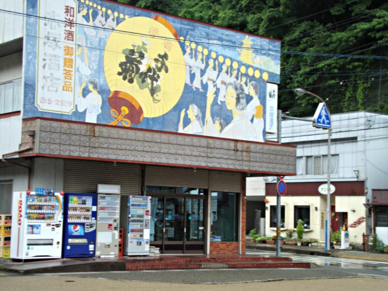Photo of a colorful painted sign above a bank of vending machines outside of Gujo Hachiman train station